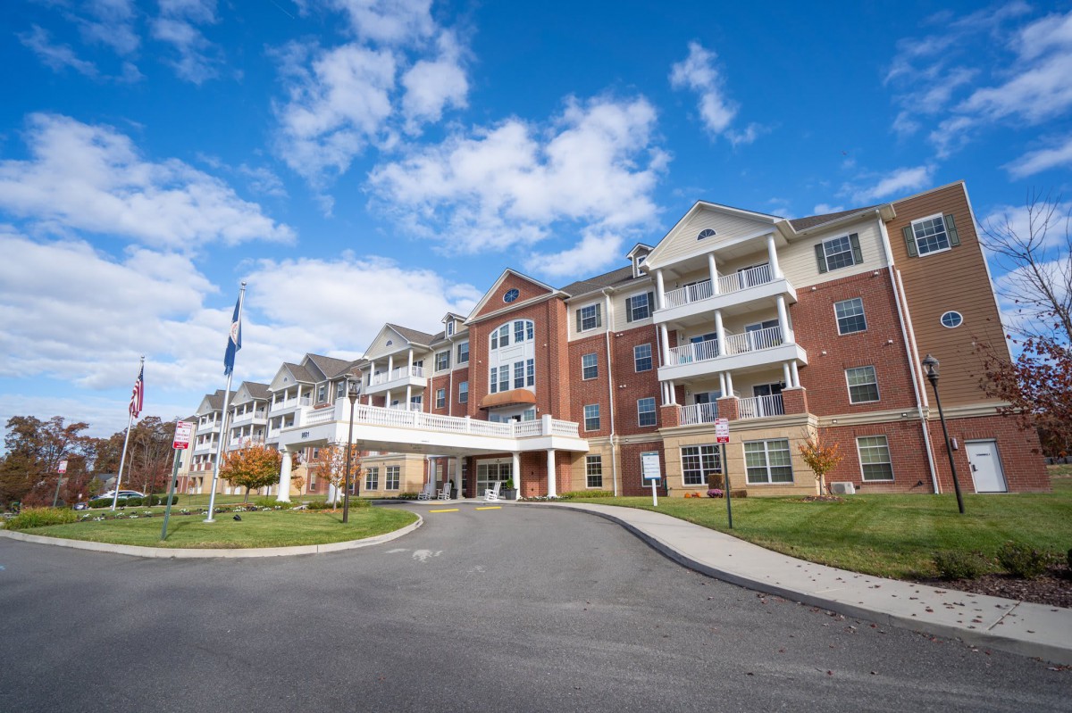 A large residential apartment building with a paved road leading to its entrance under a bright blue sky with scattered clouds.