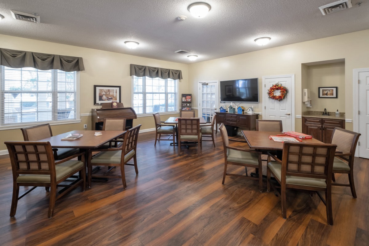 A communal dining area with wooden flooring, tables, and chairs, featuring a television on the wall.