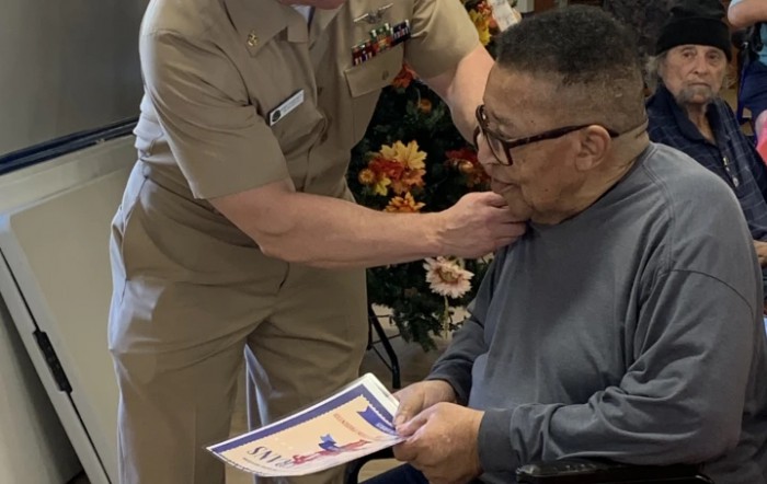 A military officer presents a medal and certificate to a seated veteran during an honoring ceremony.