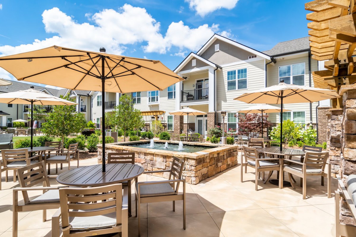 A sunny outdoor patio with tables, umbrellas, and well-maintained landscaping at an apartment complex.