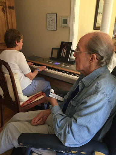 An elderly man watches as a woman plays the piano in a cozy indoor setting.