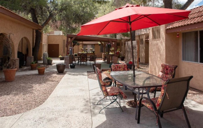 An outdoor patio area with a glass table, chairs, and a red umbrella set against a garden backdrop.
