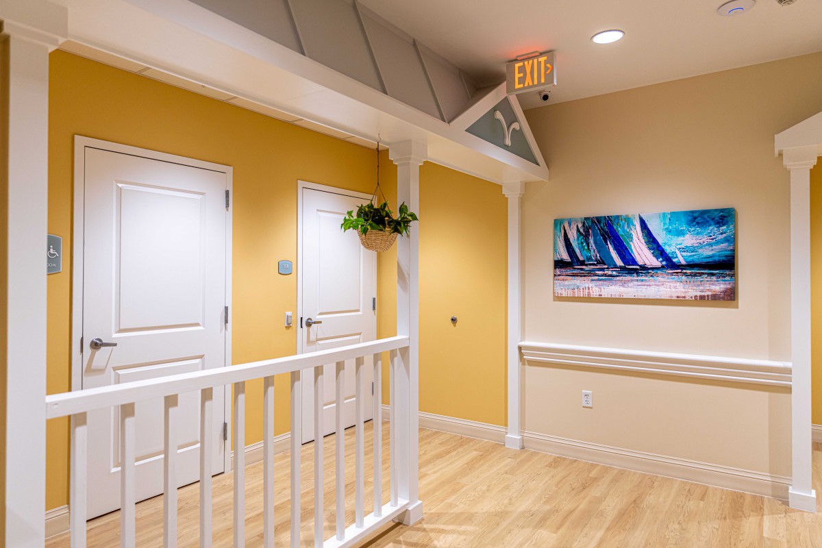 A bright hallway with yellow walls, white doors, and a wooden floor, featuring a painting and an exit sign.