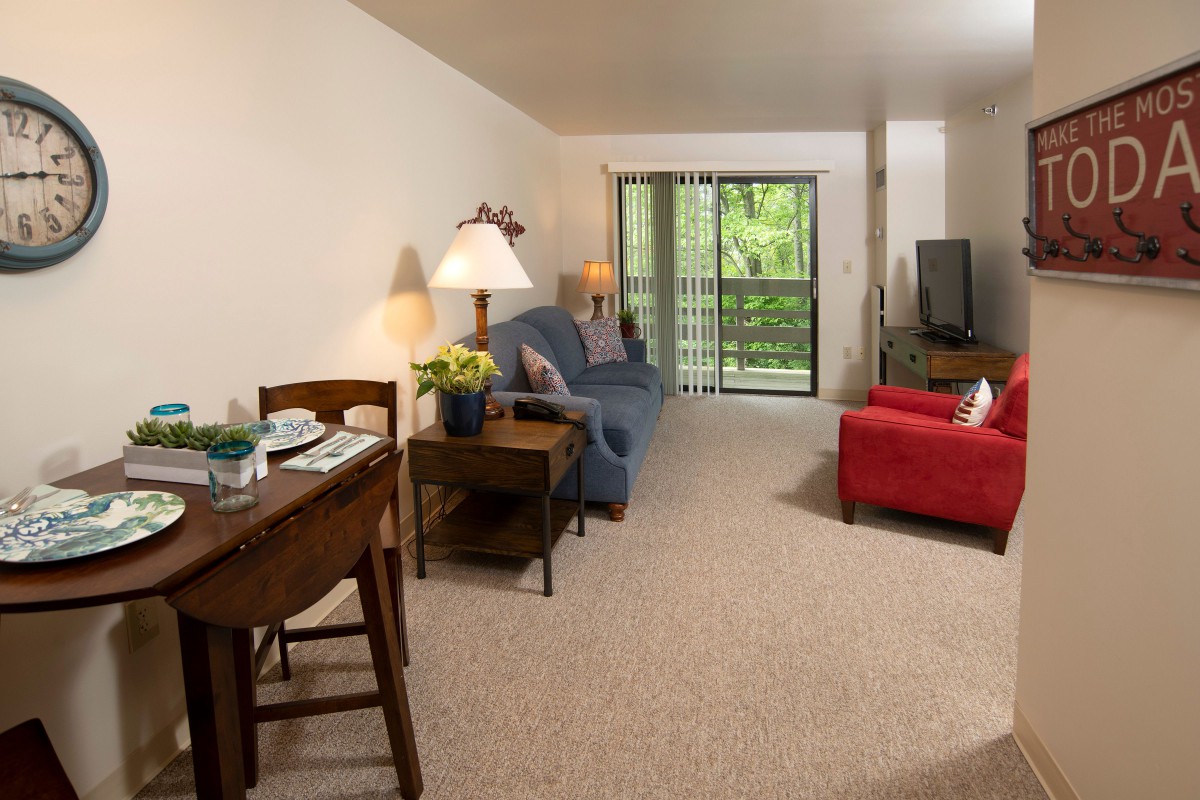 A cozy living room with a sofa, table, and television, featuring a view of greenery through sliding doors.