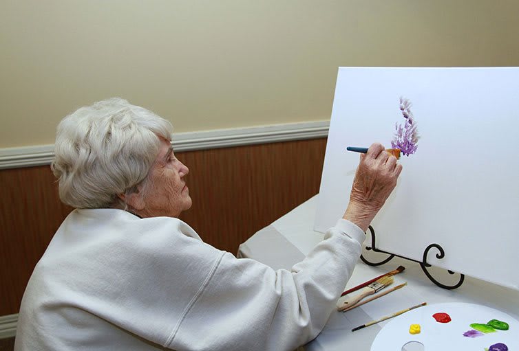An elderly woman painting on a canvas indoors.