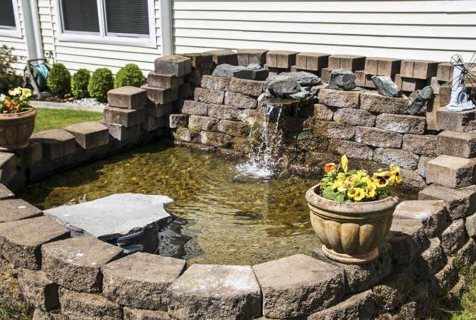 A small garden pond with a fountain and surrounding stonework, adorned with a potted plant with yellow flowers.