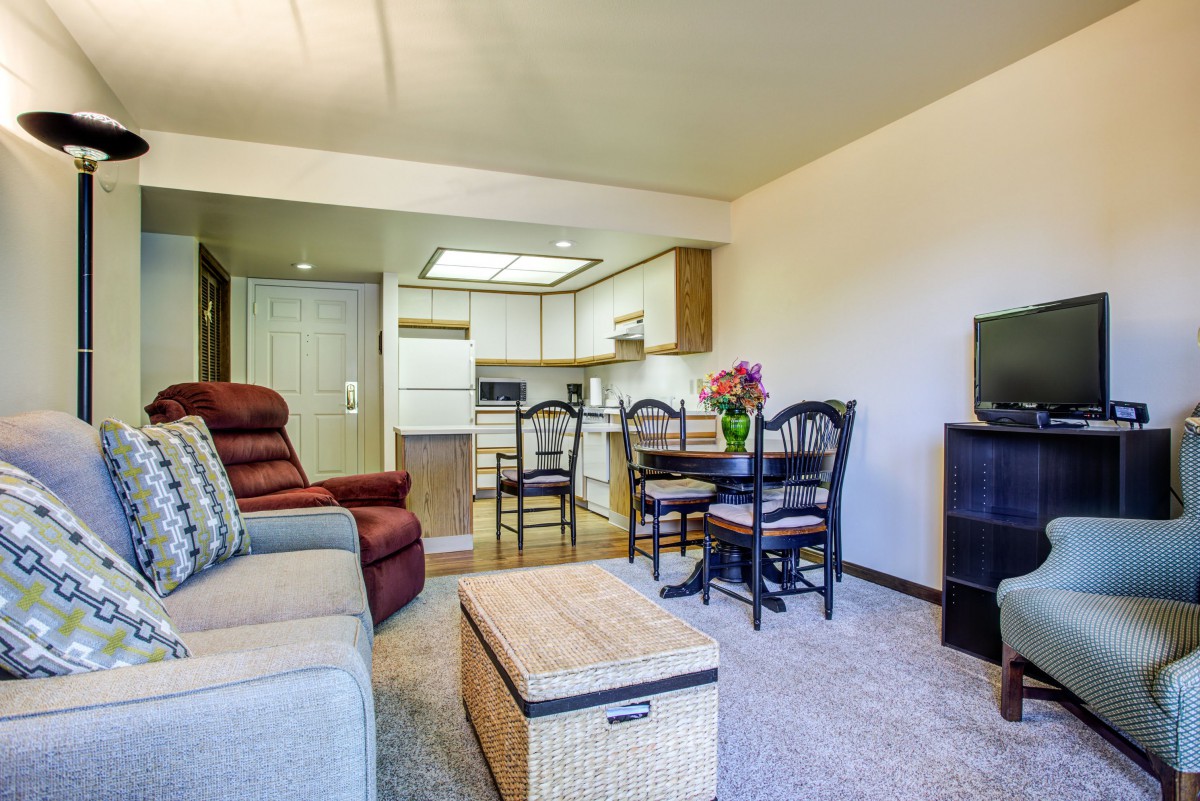 A cozy apartment living room with a sofa, chairs, and a small table, leading to an adjoining kitchen area.