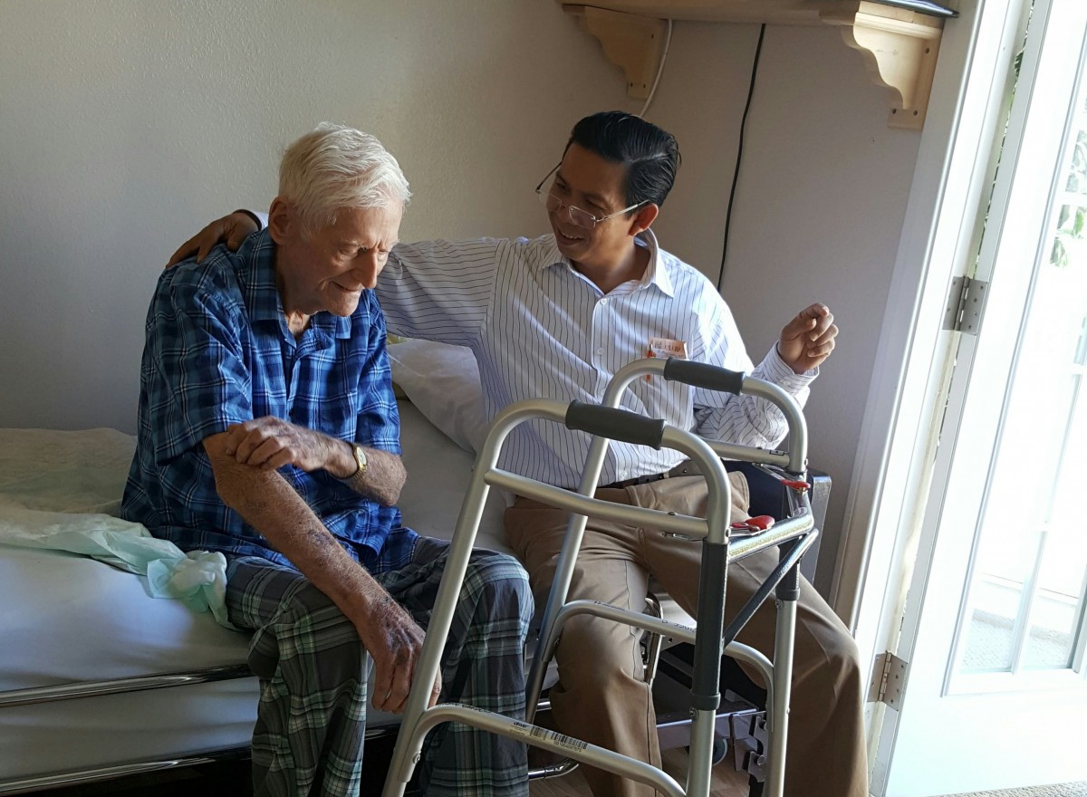 A caregiver provides support to an elderly person sitting on a bed, highlighting compassion and assistance.