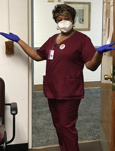 A healthcare worker in scrubs, wearing a face mask and gloves, greeting warmly.