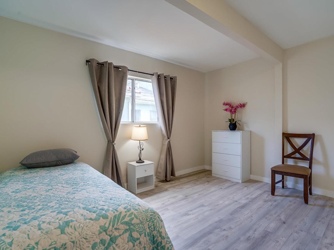 A cozy bedroom with a single bed, bedside lamp, and dresser, featuring gray curtains and light wood flooring.