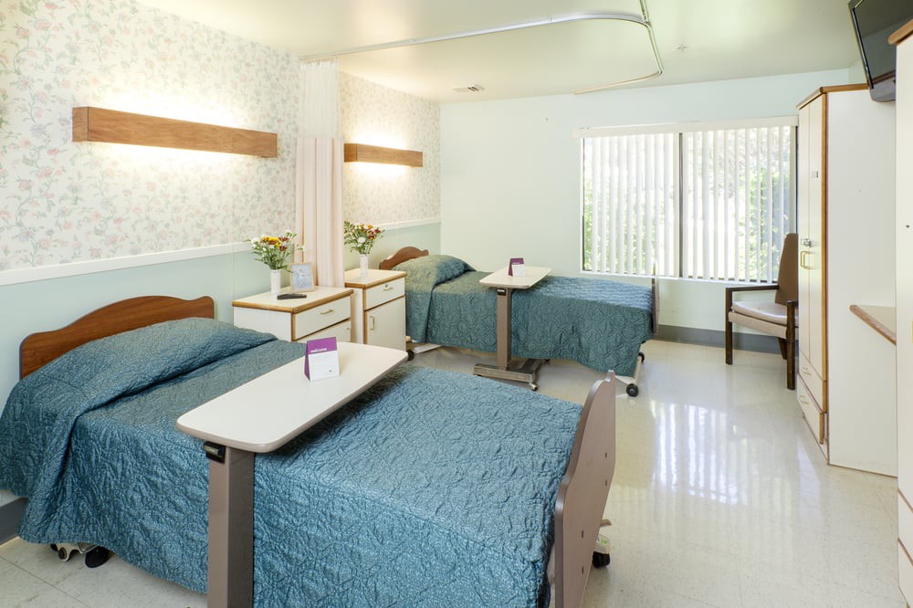 A clean and well-lit hospital room with multiple patient beds and bedside tables.