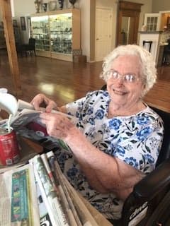 An elderly woman smiling while reading a newspaper indoors, with a soda can on the table.
