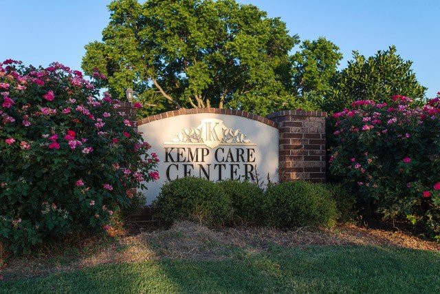 A sign for Kemp Care Center surrounded by flowers and trees.