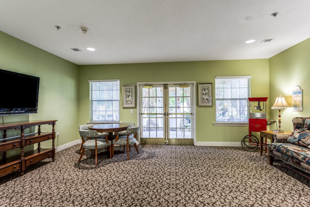 A cozy room interior featuring a table with chairs, green walls, patterned carpet, large windows, and a popcorn machine near French doors.