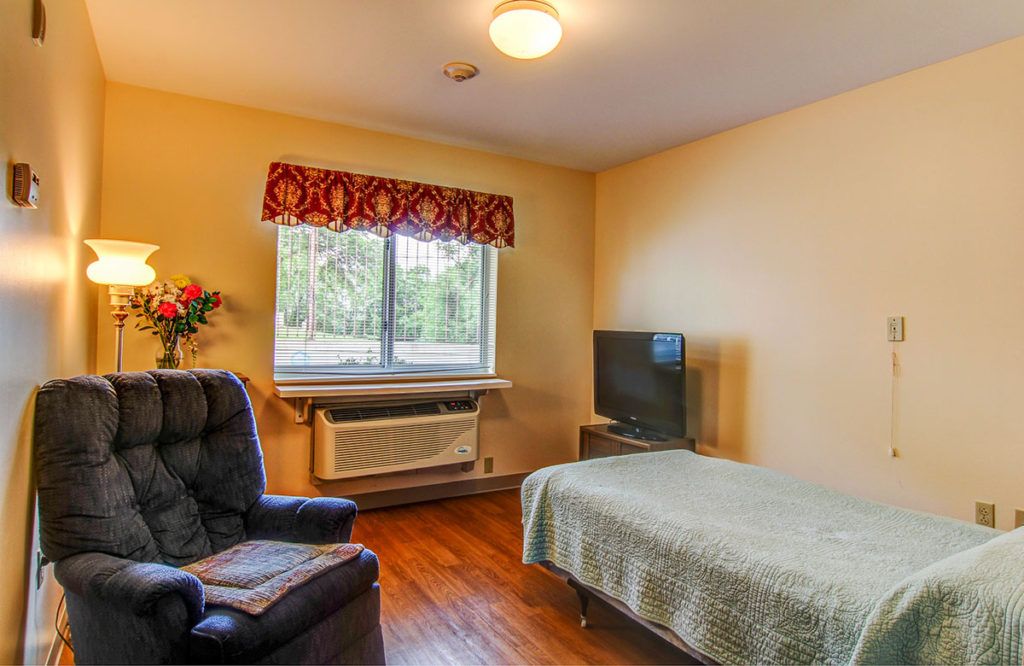 A cozy bedroom with a bed, television, and armchair, featuring a large window with decorative curtains.