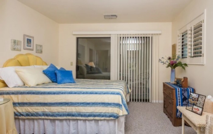 A cozy bedroom with a neatly made bed, decorative pillows, and sliding glass doors with vertical blinds.