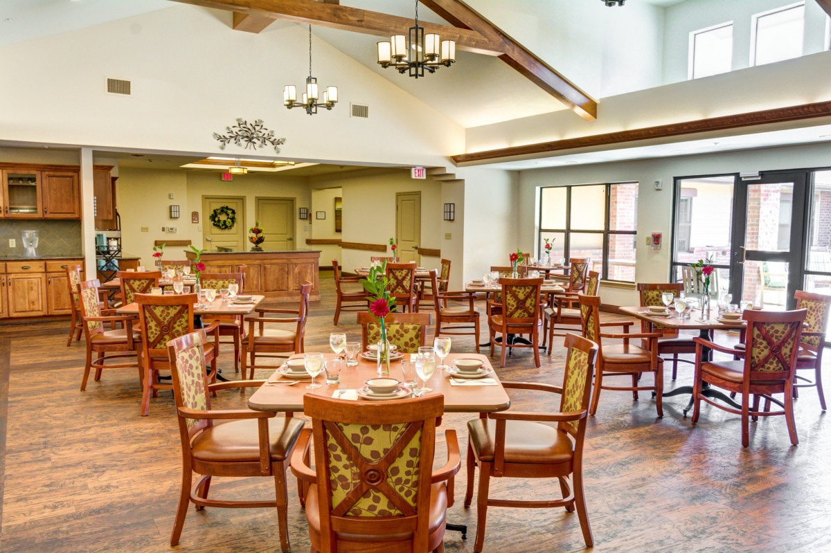 A spacious dining area with wooden tables and chairs, elegantly lit by chandeliers and natural light from large windows.