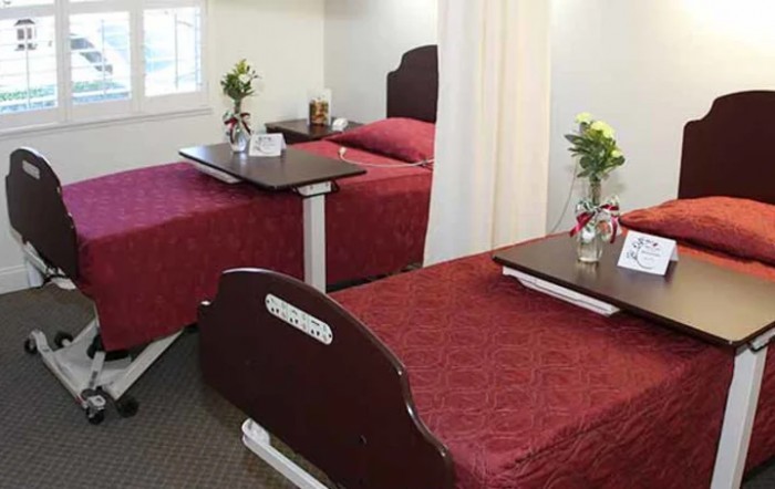 A hospital room with two patient beds featuring red bedspreads and small tables with flowers.
