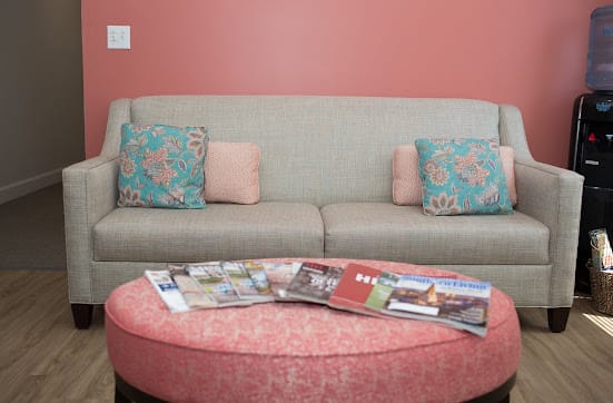 A cozy living room with a gray sofa adorned with floral cushions, a pink wall in the background, and magazines neatly arranged on a round coffee table.