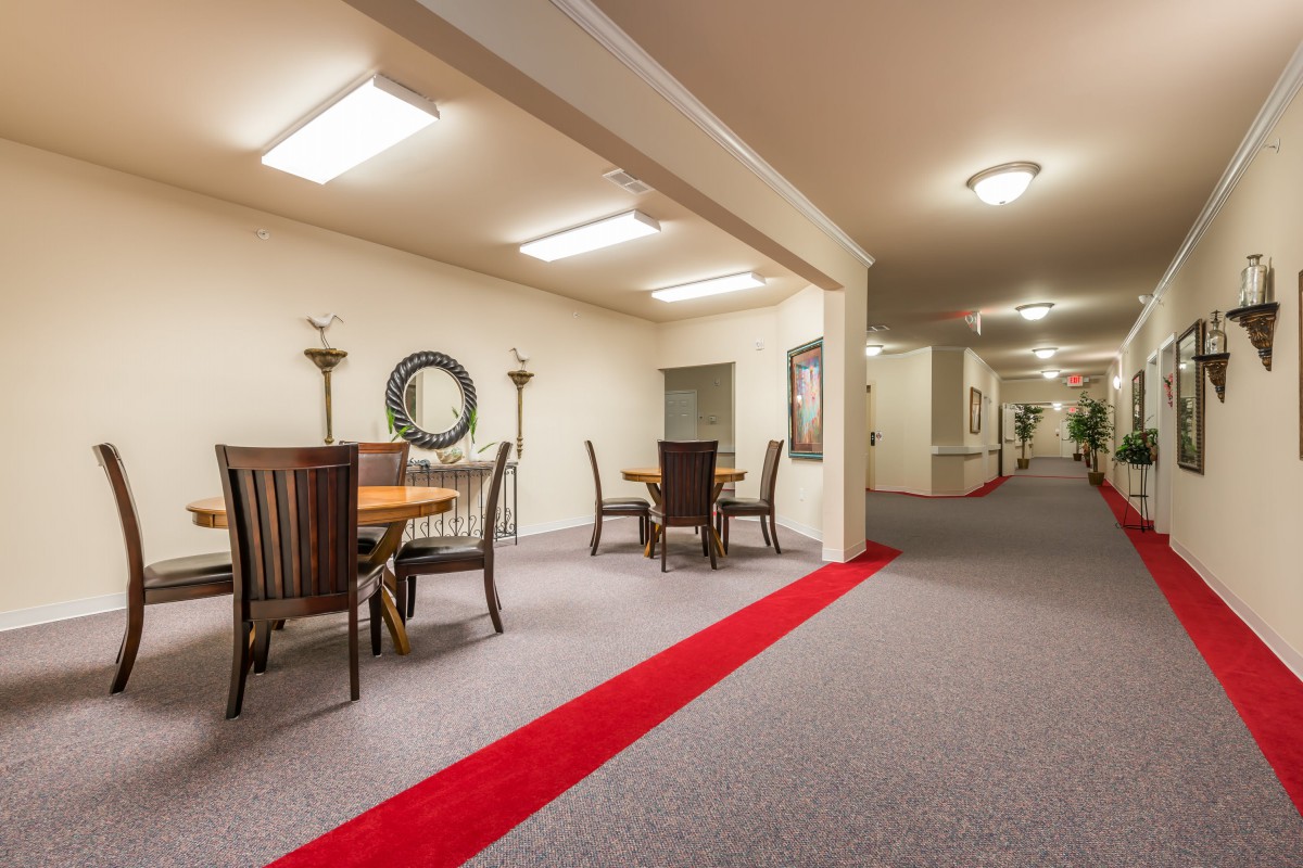 A well-lit interior space featuring a dining area with a table and chairs, adjacent to a hallway with carpet flooring.
