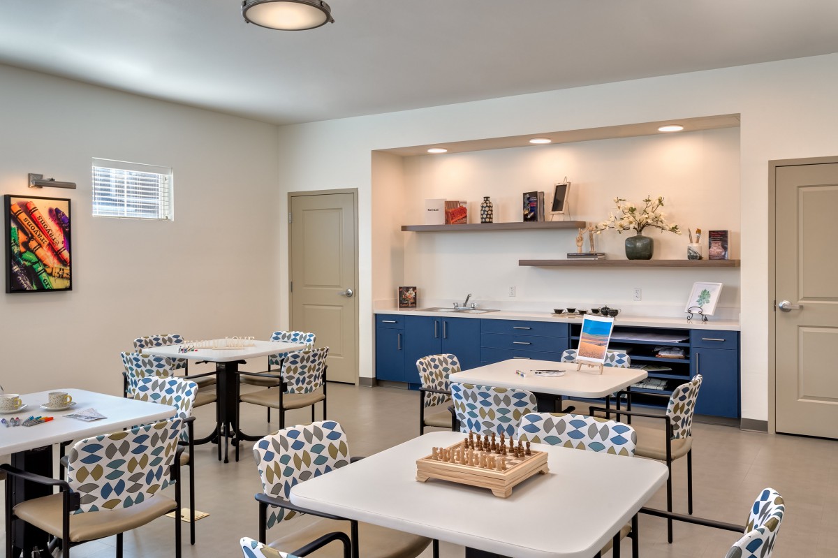 A modern community room with tables and patterned chairs, featuring a small bookshelf and decorative items.