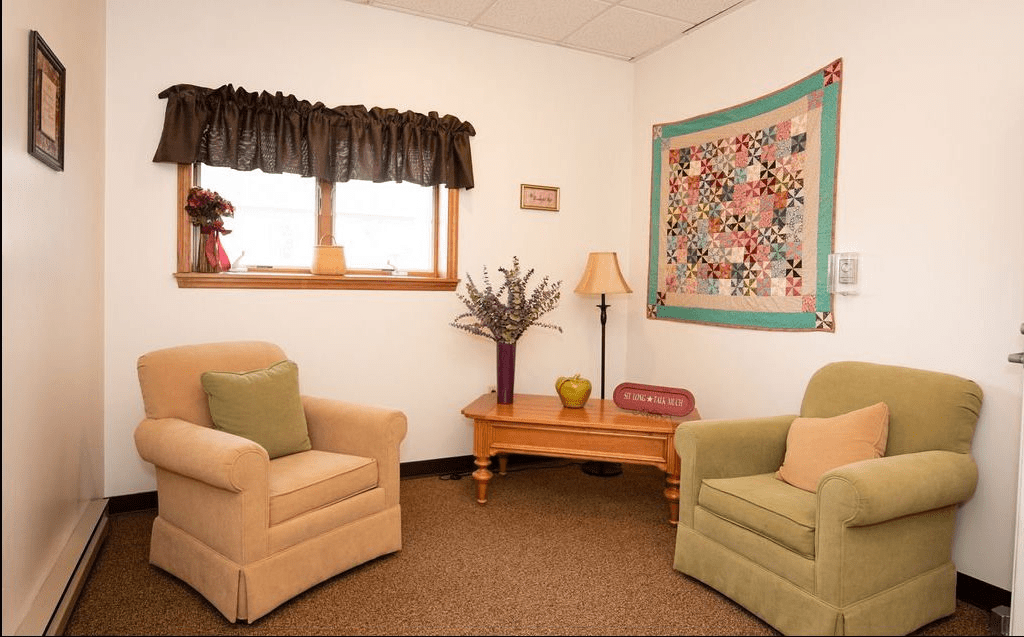 A cozy living room with two armchairs, a wooden table, and a wall-mounted quilt.