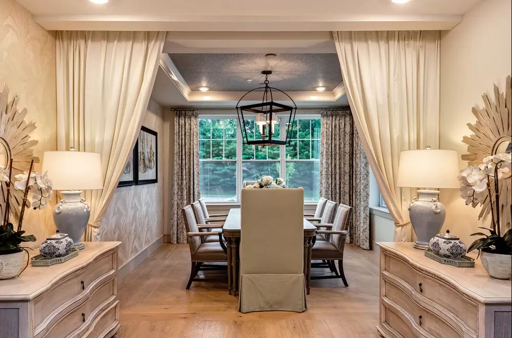 Elegant dining room with a large table, comfortable chairs, and a stylish chandelier, framed by curtains.