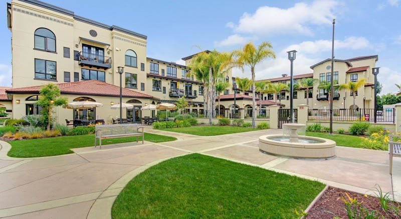 A scenic view of a courtyard with a fountain, surrounded by a multi-story building and palm trees.