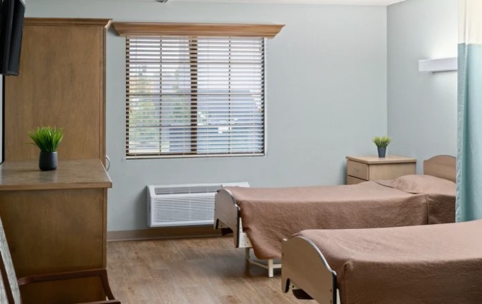 A hospital room with two beds, wooden furniture, and a window with blinds.
