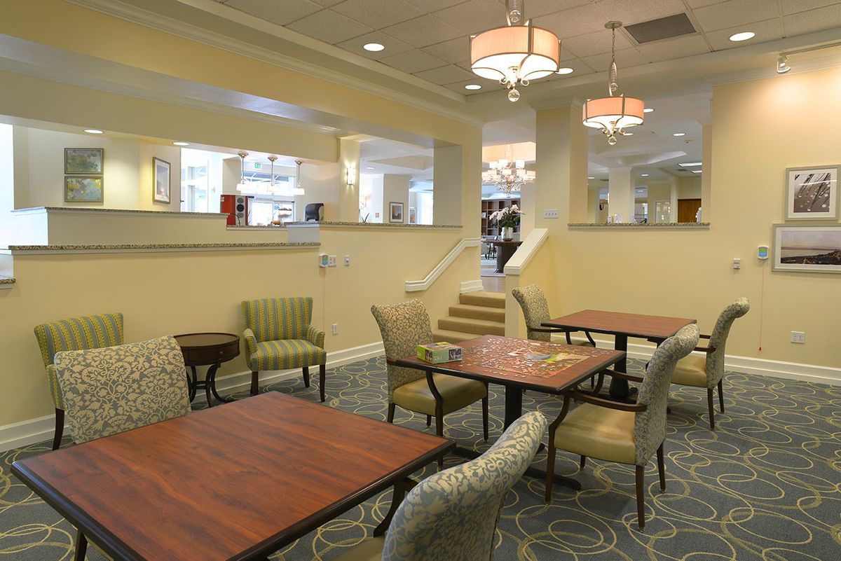 A cozy dining area with wooden tables and cushioned chairs, featuring warm lighting and a carpeted floor.