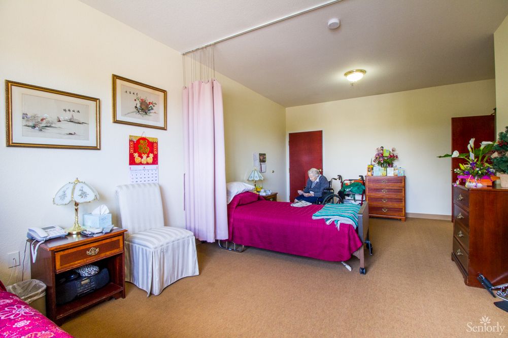 A hospital room with a single bed, where an elderly person is seated. The space includes a chair, dresser, and some wall art.