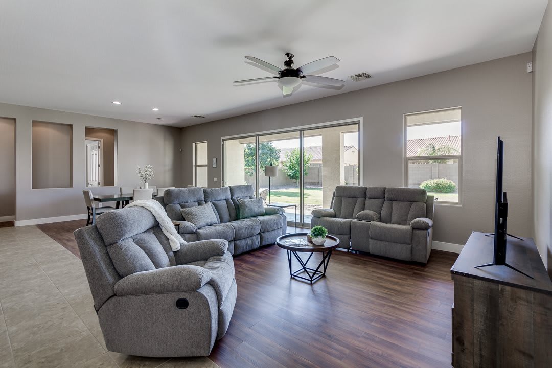 A spacious and modern living room with comfortable sofas, large windows, and a ceiling fan.