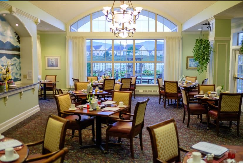 An elegant dining room with neatly arranged tables and chairs, large windows, and a chandelier.