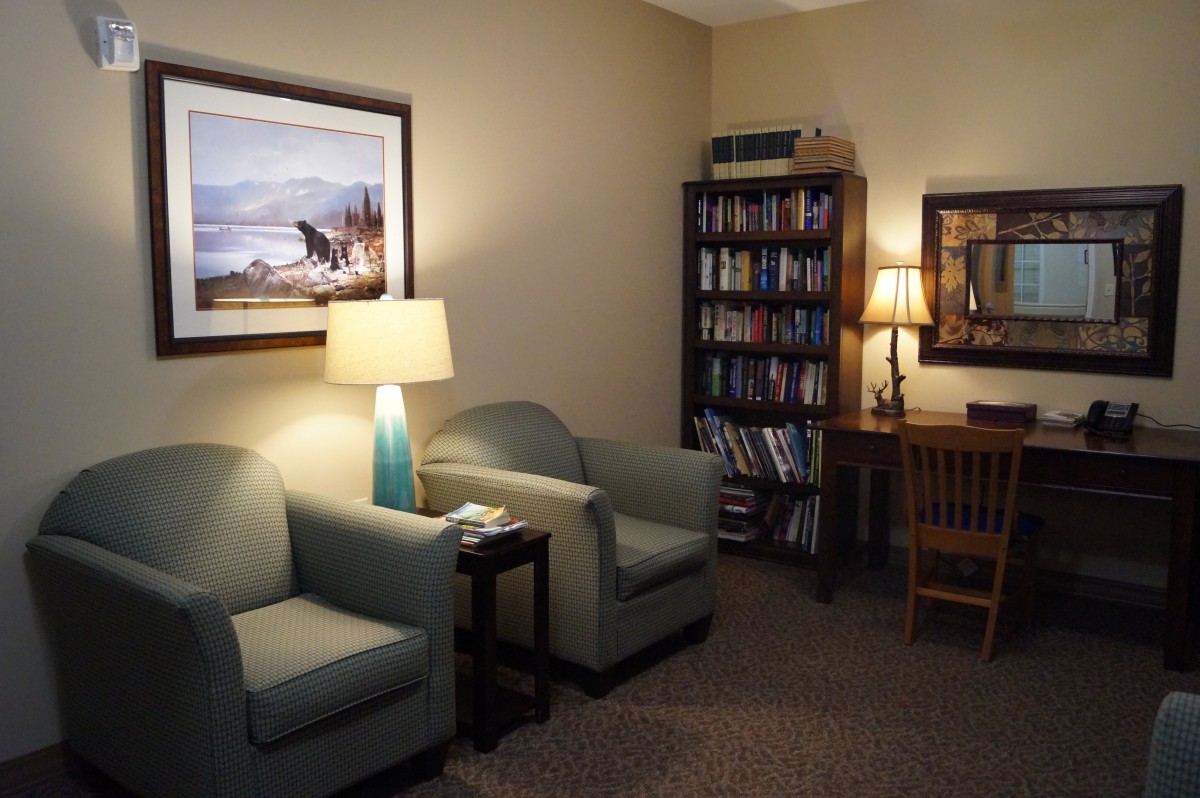 A cozy office space featuring comfortable chairs, a desk with a lamp, and a bookshelf filled with books.