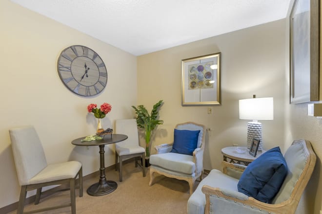 A cozy living room with elegant furnishings, including chairs, a table, and decorative elements like a wall clock and potted plant.