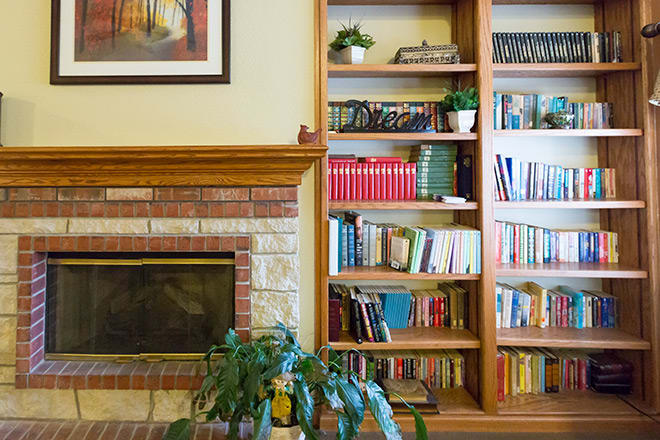 A cozy living room featuring a wooden bookshelf filled with books and potted plants, adjacent to a brick fireplace.