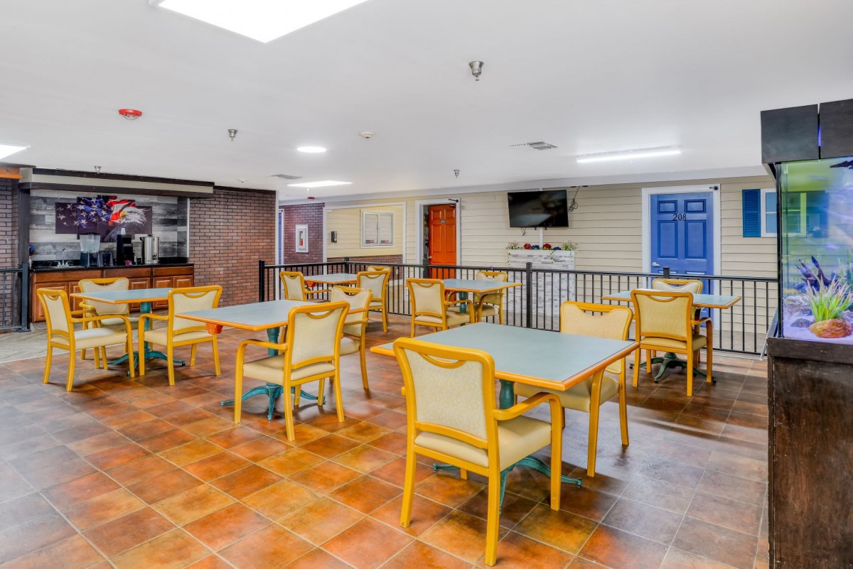 A bright indoor dining area with several tables and chairs, featuring a tiled floor and a television.