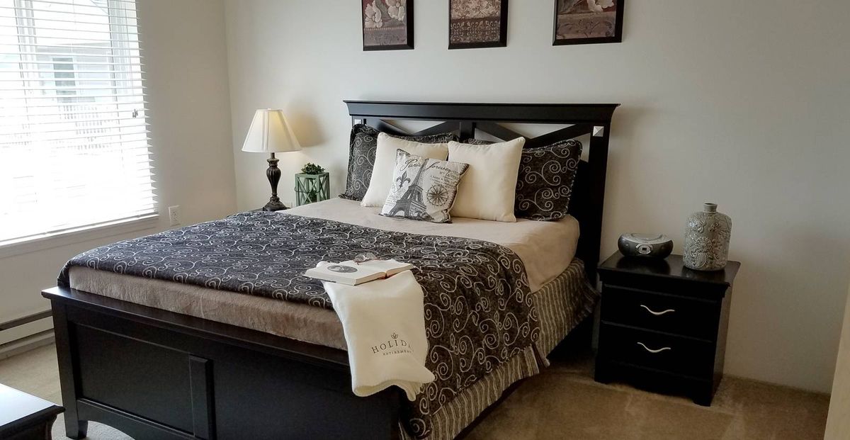 A neatly arranged bedroom with a dark wooden bedframe, patterned bedding, and decorative pillows. A lamp sits on the nightstand beside the bed.