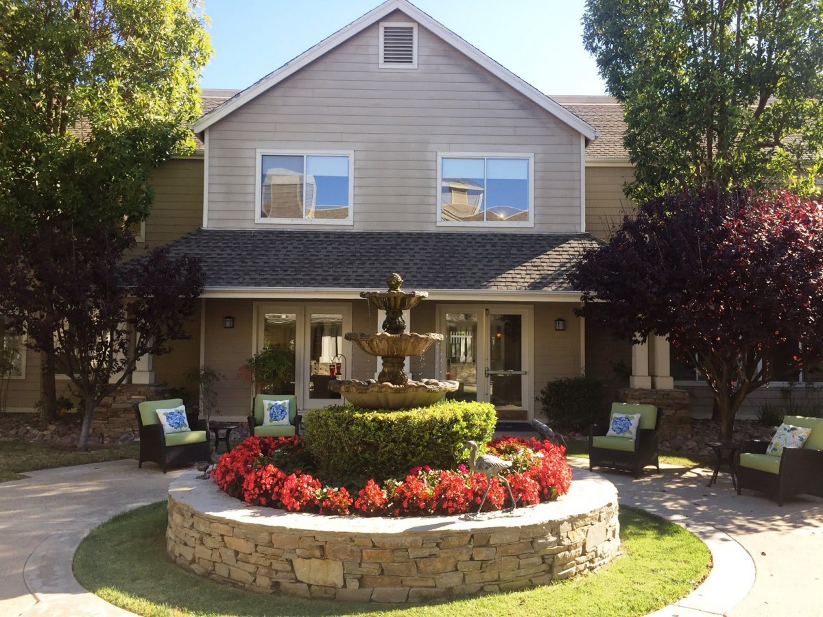 A two-story house with a beautifully landscaped garden featuring a central fountain surrounded by vibrant flowers.