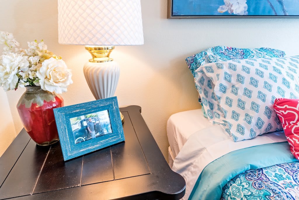 A cozy bedroom setup with a nightstand featuring a lamp, photo frame, and flowers beside a neatly made bed with patterned bedding.