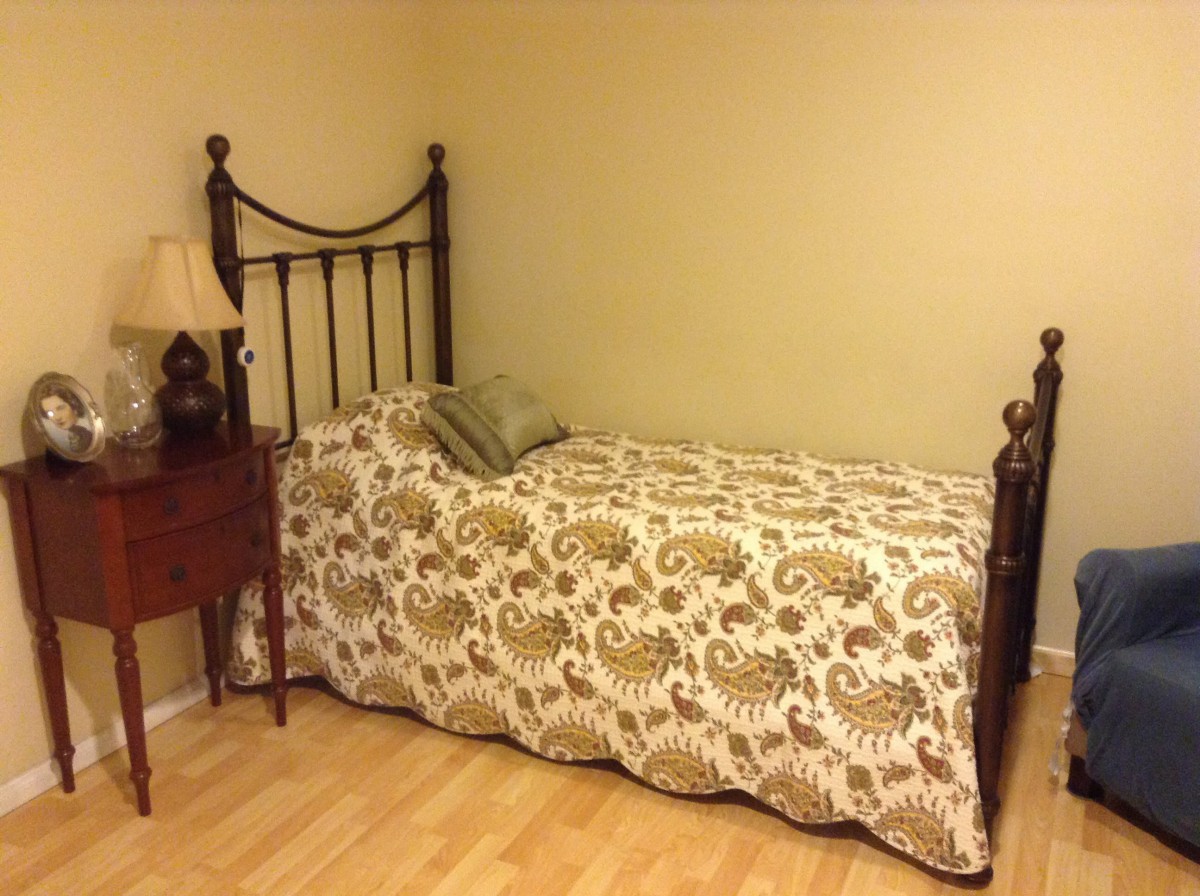 A cozy bedroom setup featuring a single bed with a patterned comforter, a decorative pillow, and a small wooden nightstand with a lamp.