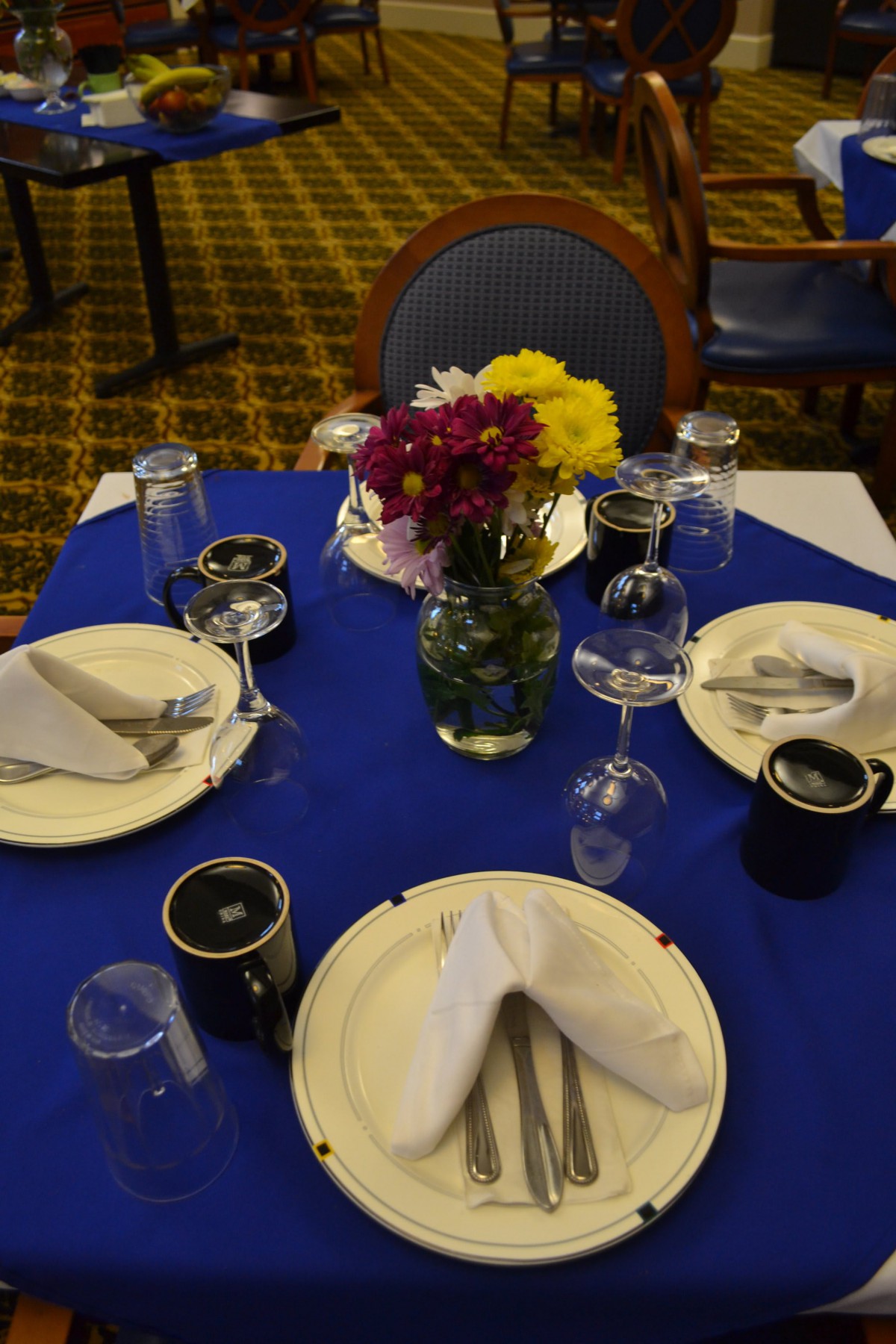 A well-arranged dining table with a blue tablecloth, featuring plates, napkins, glasses, and a floral centerpiece.