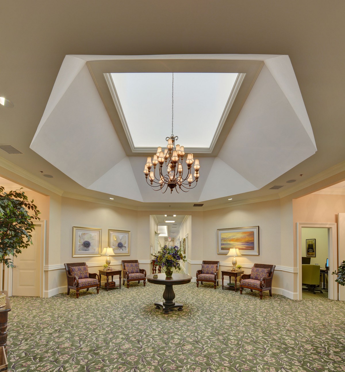 A spacious lobby with elegant decor, featuring a central chandelier and skylight, surrounded by comfortable seating and artwork.