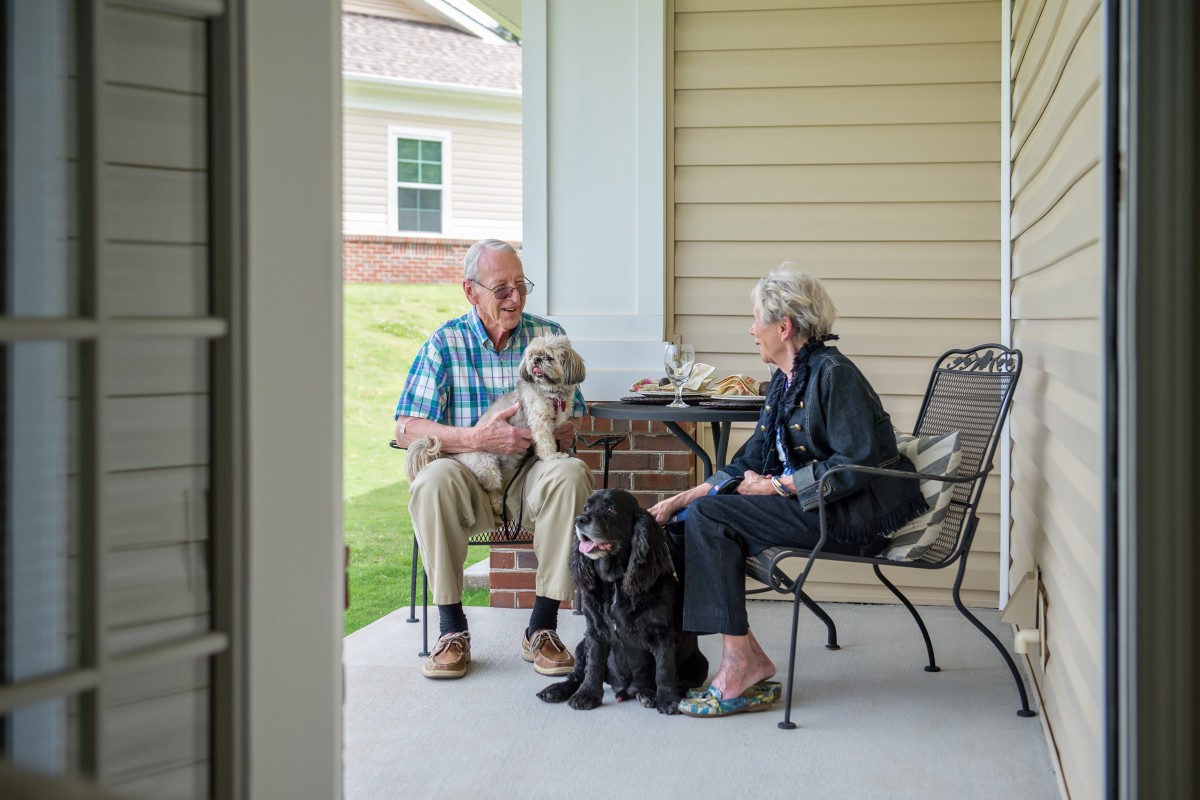 An elderly couple sitting on a porch, enjoying a conversation with their two dogs by their side.