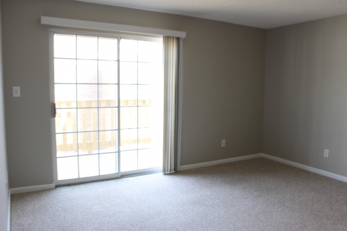 A sparsely decorated room with grey walls, featuring a sliding glass door that allows natural light to enter, and carpeted flooring.