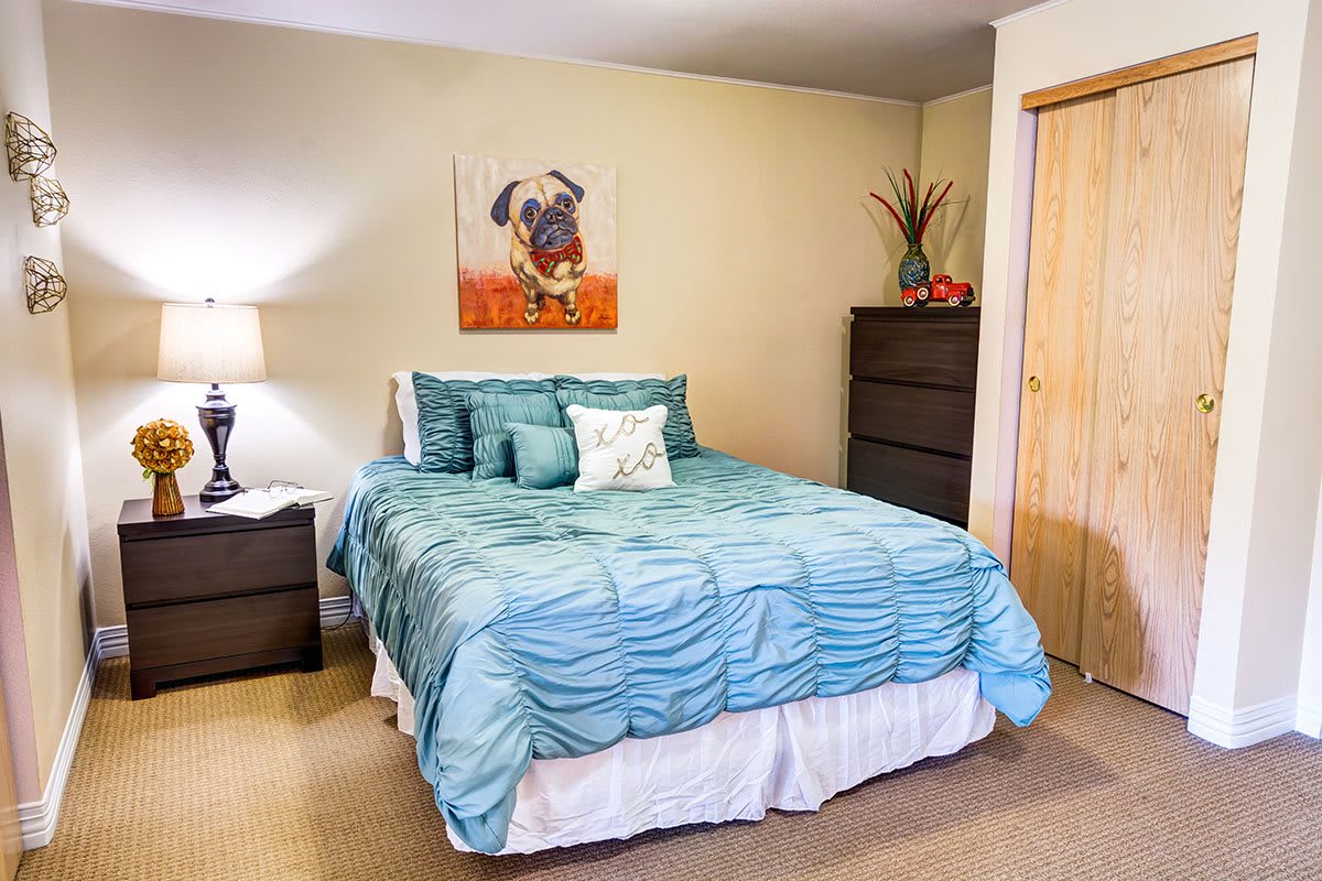 A cozy bedroom featuring a bed with a blue bedspread, a nightstand with a lamp, and wall art.