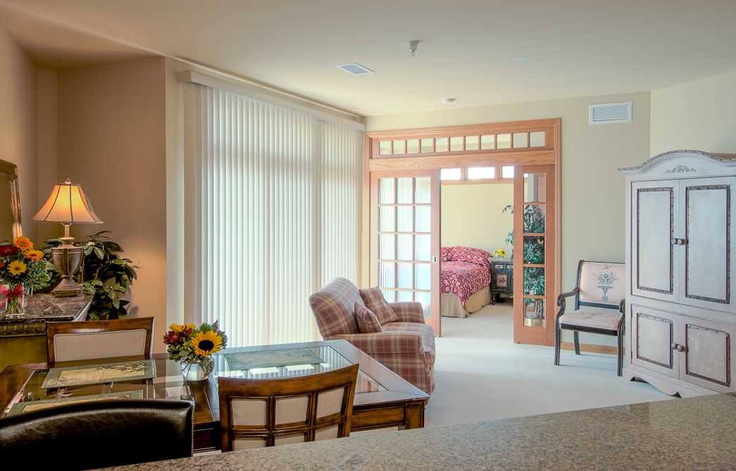 A cozy living room area with large windows, featuring comfortable furniture and a view into an adjacent bedroom.