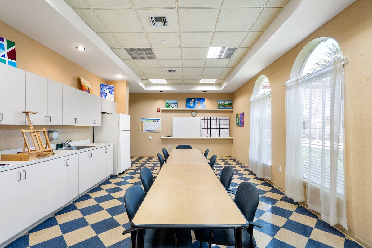 A classroom setting with a long table, surrounded by chairs, featuring a whiteboard and a kitchen area on a checkered floor.