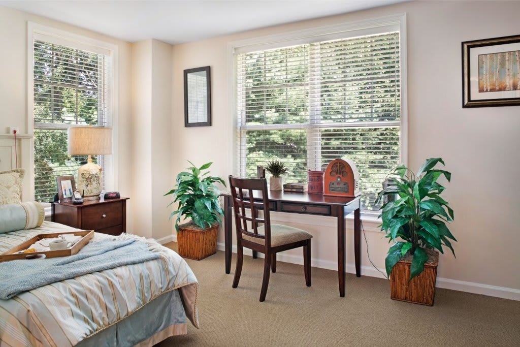 A cozy bedroom with a desk by the window, featuring natural light and green plants.