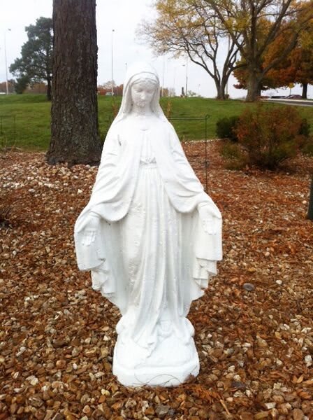A white statue of the Virgin Mary displayed outdoors in a garden setting.